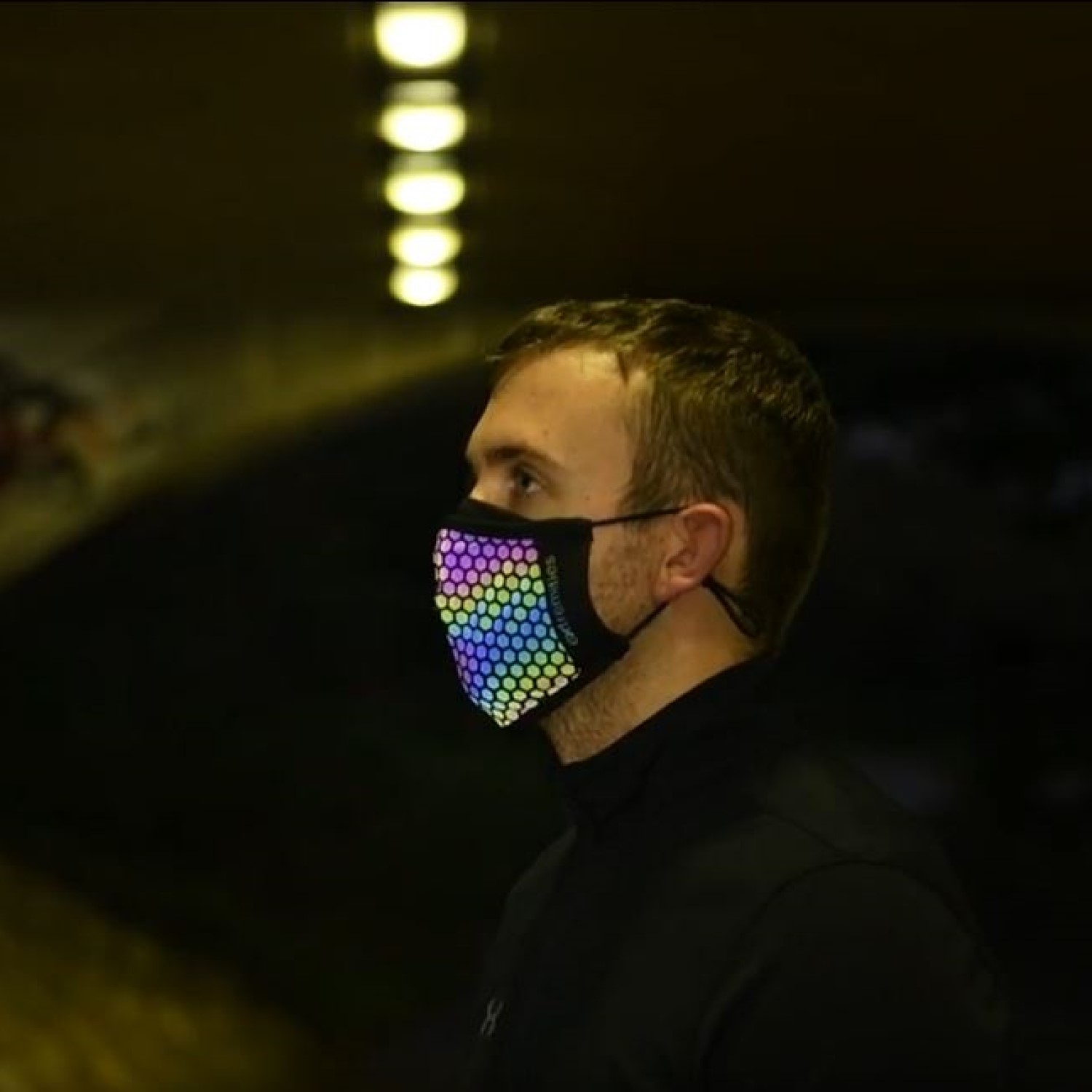 Maze-Runner-Face-Mask-Glow-1500-x1500px-1.jpg Profile of a man standing in a dim tunnel wearing a black face mask covered in a multicoloured hexagon LED pattern, with a row of glowing overhead lights.