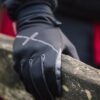 Close-up of a black insulated glove with a reflective stripe gripping a weathered wooden railing, with a blurred red coat in the background.