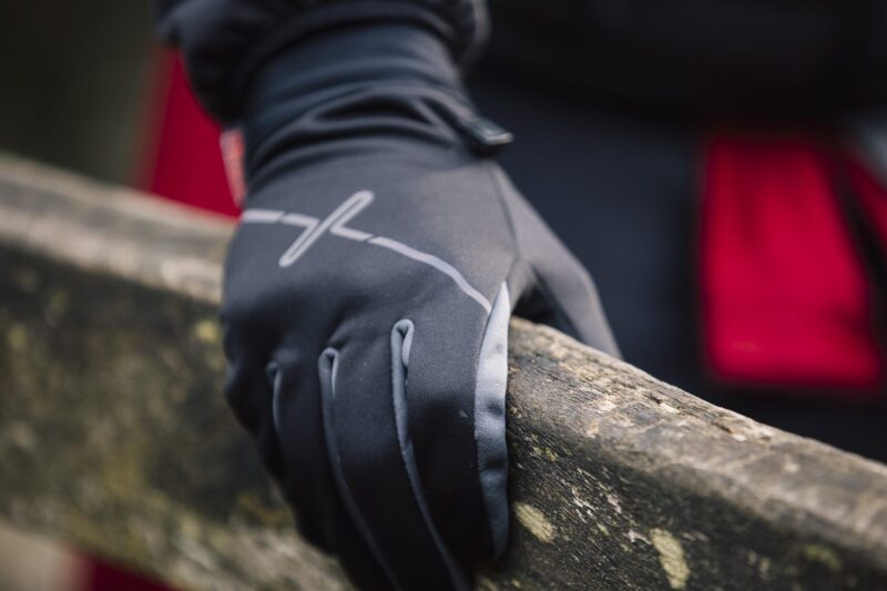 Maze-Runner-Gloves-1-3.jpg Close-up of a black insulated glove with a reflective stripe gripping a weathered wooden railing, with a blurred red coat in the background.