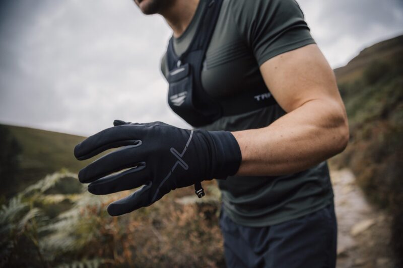 Maze-Runner-Gloves-2-3.jpg Runner on a hillside trail wearing black performance gloves and a grey-green short-sleeve top, leaning forward with a gloved hand extended toward the camera against moorland and a cloudy sky.