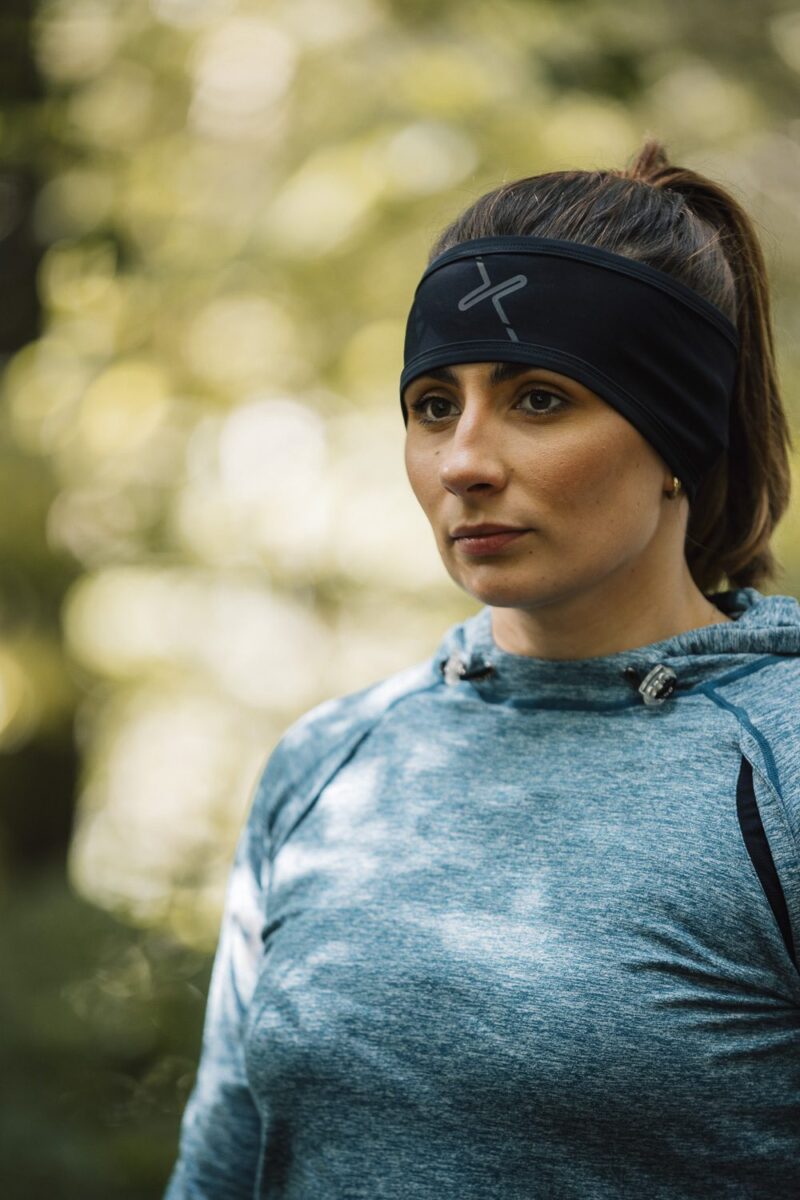 Maze-Runner-Gloves-AW25-2-3.jpg Woman with brown hair in a ponytail wearing a black headband and blue-grey athletic hoodie, standing outdoors with a neutral expression against a soft, sunlit bokeh background.
