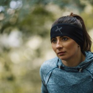 A woman with her hair in a ponytail wearing a black headband with a grey logo and a blue heathered hoodie leans forward with a focused expression against a softly blurred leafy outdoor background.