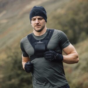 A man with a short beard runs through a misty, grassy hillside wearing a black beanie, fitted grey-green training top, a front chest vest and black gloves.