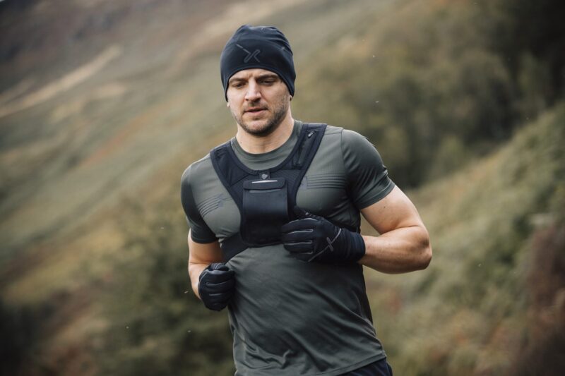 Maze-Runner-Gloves-and-Beanie-3-6.jpg A man with a short beard runs through a misty, grassy hillside wearing a black beanie, fitted grey-green training top, a front chest vest and black gloves.