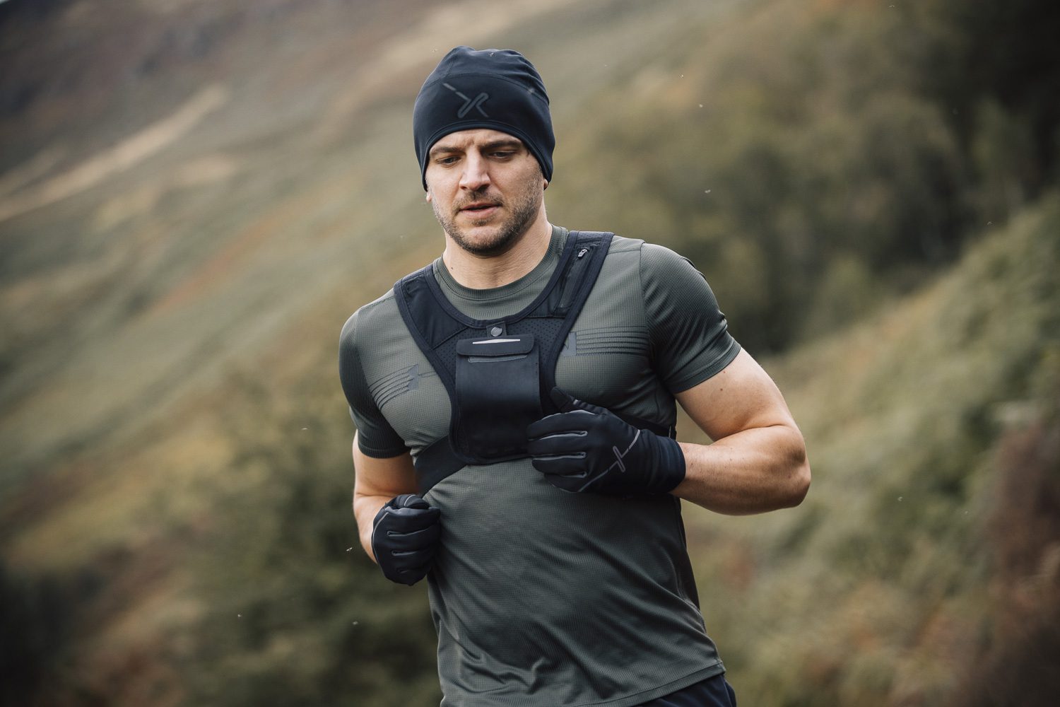 Maze-Runner-Gloves-and-Beanie-3-6.jpg A man with a short beard runs through a misty, grassy hillside wearing a black beanie, fitted grey-green training top, a front chest vest and black gloves.