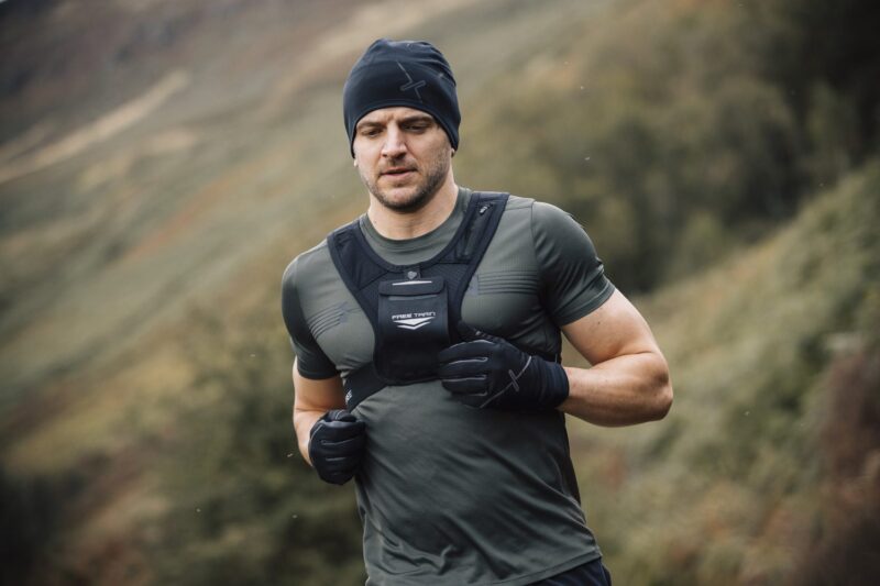Maze-Runner-Gloves-and-Beanie-3-7.jpg Man wearing a black beanie, fitted green training shirt, chest-weighted vest labelled 'FREE TRAIN' and black gloves running outdoors on a blurred hillside landscape.