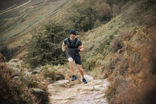 Man in a beanie, gloves and a weighted vest running uphill mid-stride on a rocky country trail through ferns and rolling grassy hills.