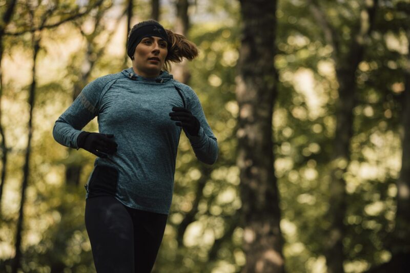 Maze-Runner-Headband-and-Gloves-AW25-7-3.jpg Woman jogging through a leafy forest wearing a teal long-sleeve top, black headband and gloves, her hair swept back with trees softly blurred behind her