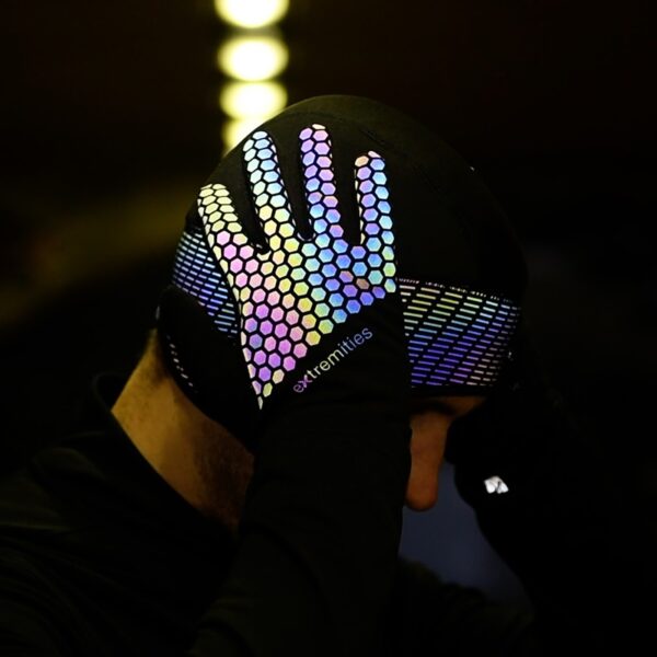 Maze-Runner-glove-and-took-lifestyle-in-tunnel-new-print-glove-1500x1500px-1.jpg Person wearing a black beanie with a hand pressed to the side of their head, the glove covered in multicoloured reflective hexagonal grip pattern and a diagonal-striped reflective band, with the extremities logo on the wrist.