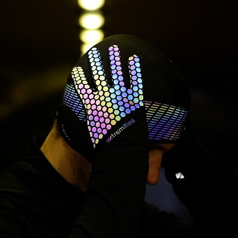 Person wearing a black beanie with a hand pressed to the side of their head, the glove covered in multicoloured reflective hexagonal grip pattern and a diagonal-striped reflective band, with the extremities logo on the wrist.