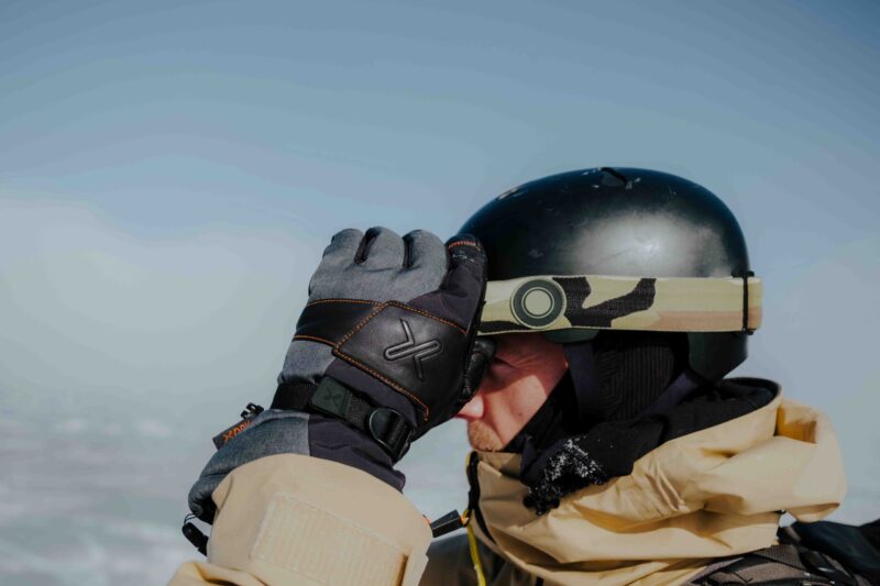 Meridian-3-scaled-4.jpg Close-up of a person in a beige winter jacket wearing a black helmet with a camouflage goggle strap, lifting a grey-and-black glove to adjust goggles against a pale blue sky and snowy background.