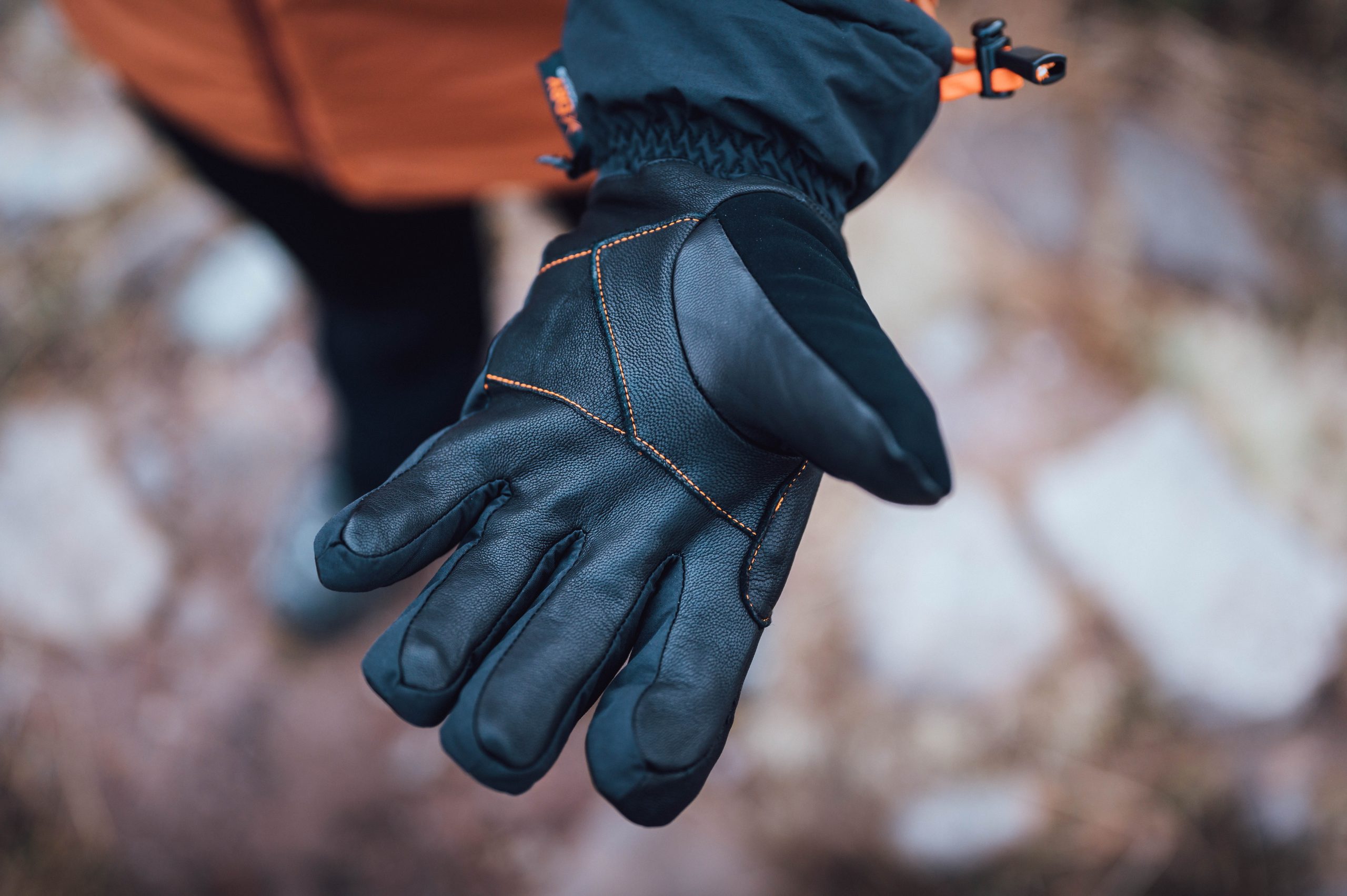 Close-up of an outstretched black leather winter glove with orange stitching and an insulated cuff with a drawcord, held against a blurred rocky outdoor background.