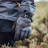 Hand wearing a grey-and-black insulated glove with orange stitching, an embossed X logo and an adjustable wrist strap, hanging by the side of a person in a dark jacket against a blurred heather-covered outdoor background.