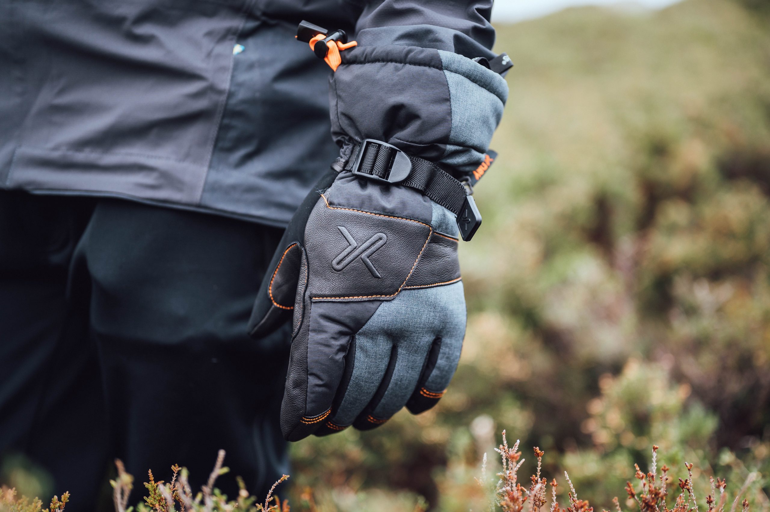 Hand wearing a grey-and-black insulated glove with orange stitching, an embossed X logo and an adjustable wrist strap, hanging by the side of a person in a dark jacket against a blurred heather-covered outdoor background.