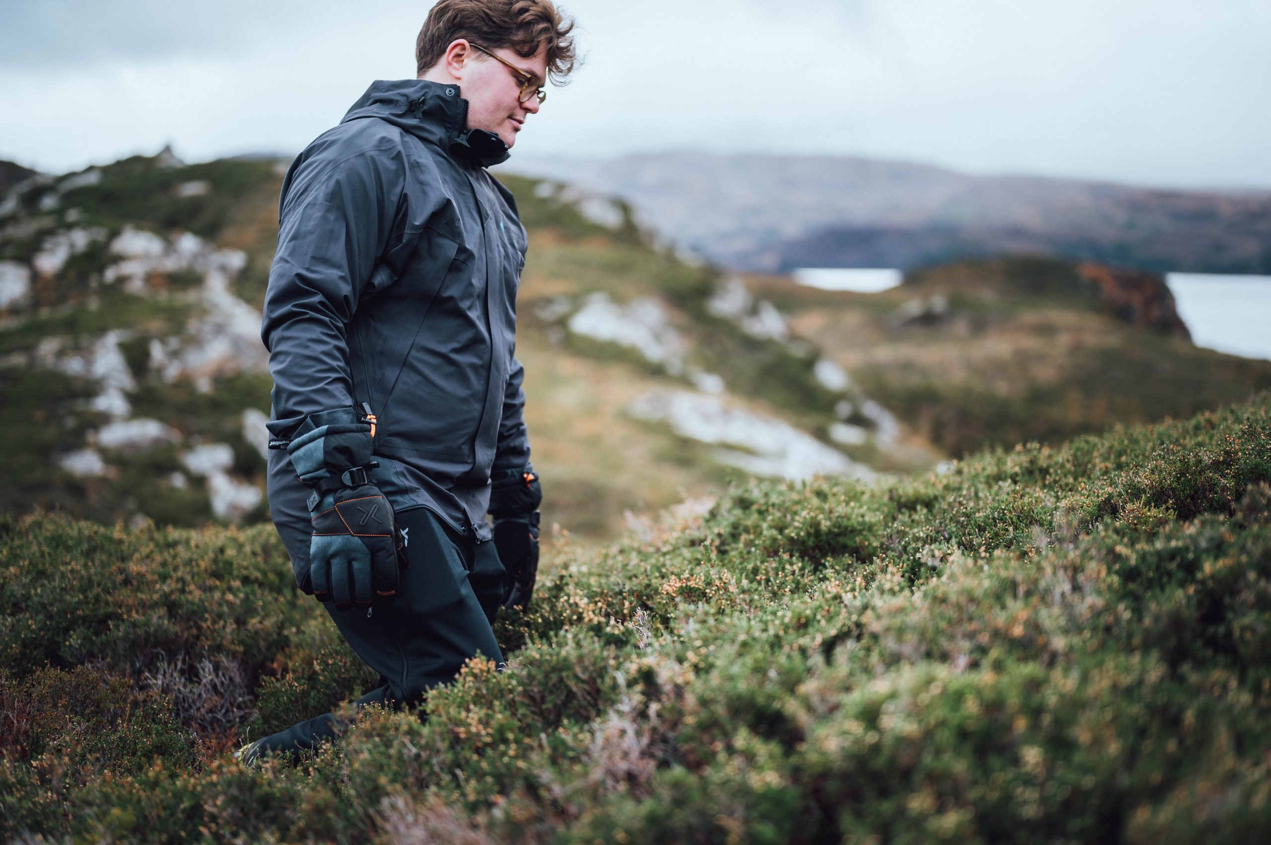 A person wearing a dark waterproof jacket, gloves and glasses walks through low green heath and rocky outcrops with a distant loch under an overcast sky.