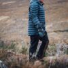 Person wearing a blue puffer jacket, mittens and dark trousers walking across windswept moorland with brown grasses and low shrubs.