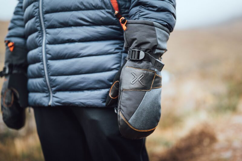 Close-up of a person wearing a blue‑grey puffer jacket and grey insulated mittens with black leather patches, orange stitching and a buckle strap, standing outdoors against a blurred brown landscape.