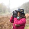 Merlin-Highclere-scaled-2.jpg Person in a black hat decorated with a feather and a red jacket, wearing gloves and a scarf, looking through binoculars in light rain over an autumn field.
