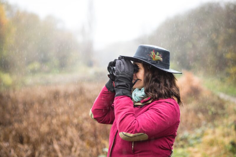 Merlin-Highclere-scaled-2.jpg Person in a black hat decorated with a feather and a red jacket, wearing gloves and a scarf, looking through binoculars in light rain over an autumn field.
