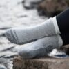 Mountain-Toester-26MTO-3000x3000px-300dpi-scaled-4.jpg Feet in thick light-grey ribbed socks with dark-grey reinforced toes and heels, legs in black leggings, resting on a rock beside a blurred flowing stream.