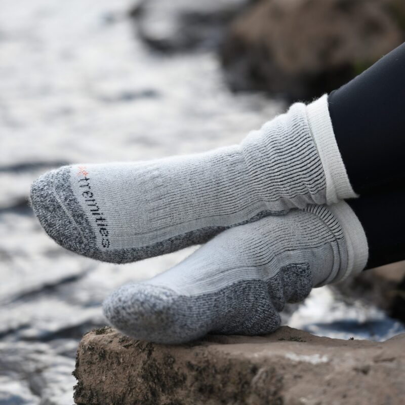 Mountain-Toester-26MTO-3000x3000px-300dpi-scaled-4.jpg Feet in thick light-grey ribbed socks with dark-grey reinforced toes and heels, legs in black leggings, resting on a rock beside a blurred flowing stream.