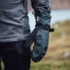Mountain_Mitt_LS-11-scaled-4.jpg Close-up of a person's side wearing a dark waterproof jacket and a black Gore-Tex mitten with an adjustable wrist strap, set against a blurred outdoor landscape.