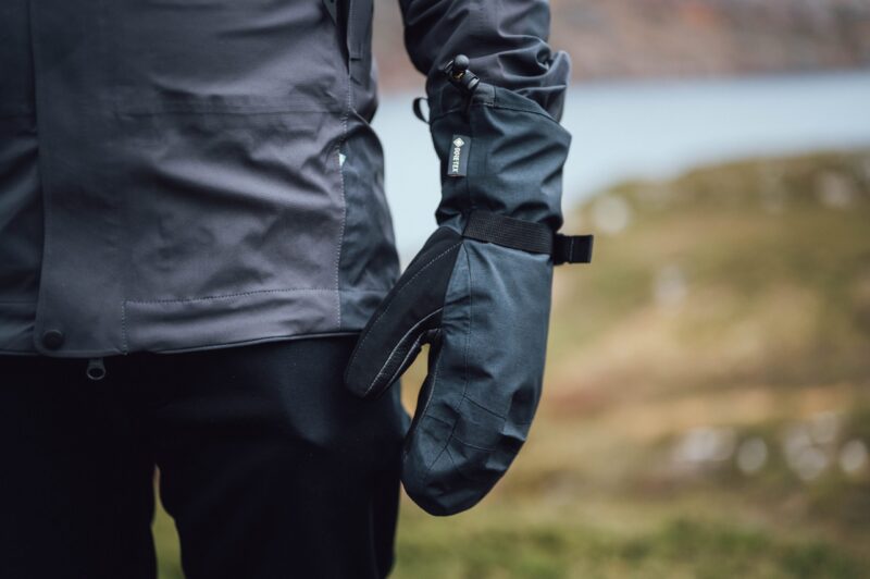 Mountain_Mitt_LS-11-scaled-4.jpg Close-up of a person's side wearing a dark waterproof jacket and a black Gore-Tex mitten with an adjustable wrist strap, set against a blurred outdoor landscape.