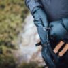 Mountain_Mitt_LS-2-scaled-4.jpg Close-up of a person wearing dark puffy mittens adjusting a strap on a black hand cover while holding hiking poles with cork handles against a blurred green ground background.