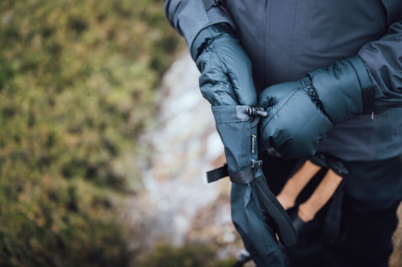 Mountain_Mitt_LS-2-scaled-4.jpg Close-up of a person wearing dark puffy mittens adjusting a strap on a black hand cover while holding hiking poles with cork handles against a blurred green ground background.