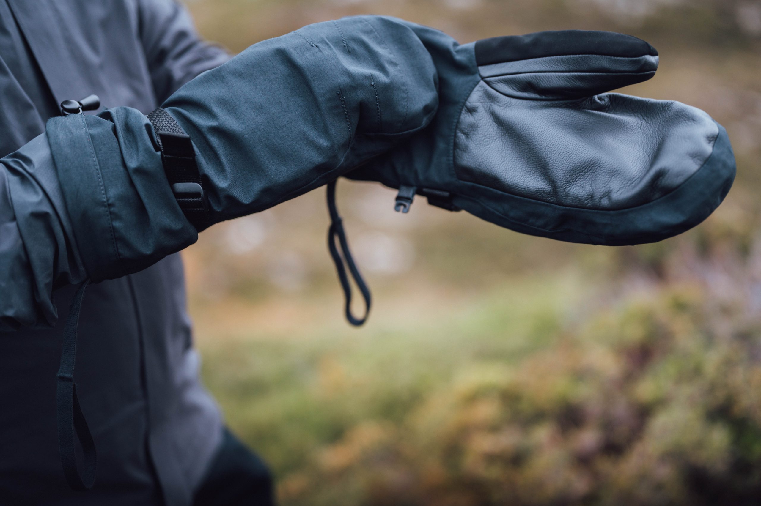 A person wearing dark waterproof mittens with leather palms and adjustable wrist straps, hands extended against a blurred grassy outdoor background.