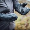 Mountain_Mitt_LS-6-scaled-4.jpg Close-up of a person wearing black heavy-duty mitten-style waterproof gloves with leather palms and adjustable wrist straps, held out against a blurred outdoor landscape