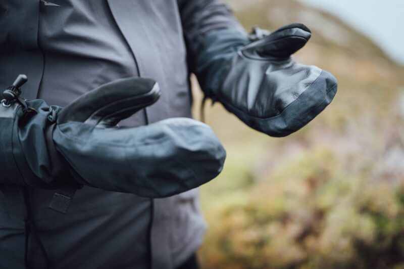 Mountain_Mitt_LS-6-scaled-4.jpg Close-up of a person wearing black heavy-duty mitten-style waterproof gloves with leather palms and adjustable wrist straps, held out against a blurred outdoor landscape