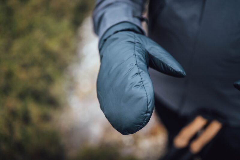 Mountain_Mitt_LS-scaled-4.jpg Close-up of an outstretched padded teal mitten showing visible stitching, worn with a dark jacket against a blurred outdoor background.