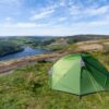 Panacea-2-3-1.jpg A bright green camping tent pitched on a grassy hillside overlooking a winding reservoir with a dam, surrounded by wooded valleys and rolling green hills beneath a blue sky streaked with wispy clouds.