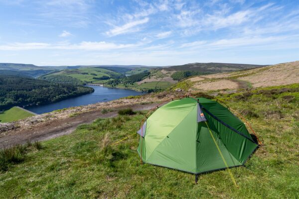 Panacea-2-3-1.jpg A bright green camping tent pitched on a grassy hillside overlooking a winding reservoir with a dam, surrounded by wooded valleys and rolling green hills beneath a blue sky streaked with wispy clouds.