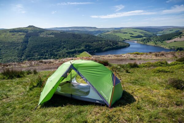 Panacea-3-2-With-Logo-3.jpg Bright green two-person tent pitched on a grassy hilltop overlooking a forested valley and a blue reservoir with an arched bridge and rolling green hills under a clear sky.