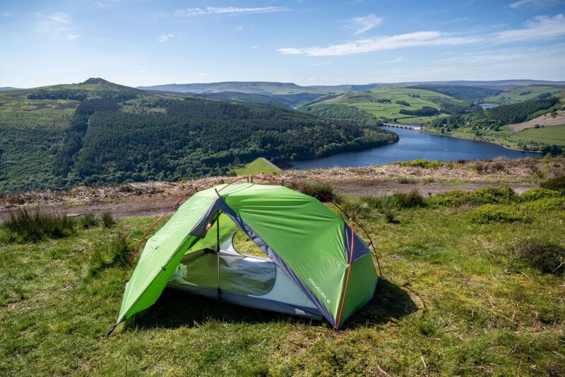 Panacea-3-2-With-Logo-3.jpg Bright green two-person tent pitched on a grassy hilltop overlooking a forested valley and a blue reservoir with an arched bridge and rolling green hills under a clear sky.