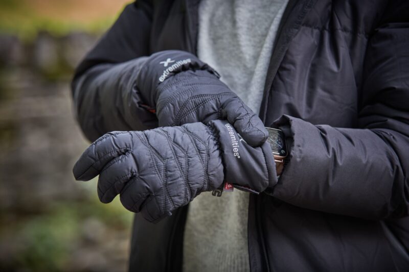 Paradox-Glove-scaled-4.jpg Close-up of a person outdoors wearing quilted black 'extremities' insulated gloves and a dark puffer jacket, one glove adjusting a smartwatch on the other wrist against a blurred stone-wall background.