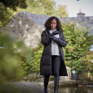Woman in a long black puffer coat, grey sweater and gloves adjusts her sleeve while walking along a path by a stream, with a stone cottage and autumnal greenery in the background.