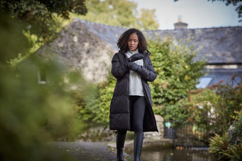 Paradox-Mitt-scaled-4.jpg Woman in a long black puffer coat, grey sweater and gloves adjusts her sleeve while walking along a path by a stream, with a stone cottage and autumnal greenery in the background.
