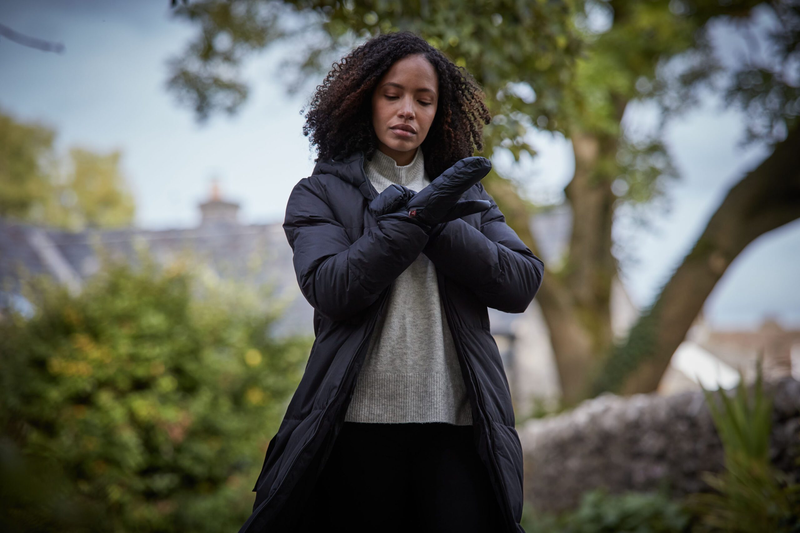 Woman with curly hair standing outdoors in a long black puffer coat and grey sweater, putting on black insulated mittens with trees and a stone wall blurred in the background.