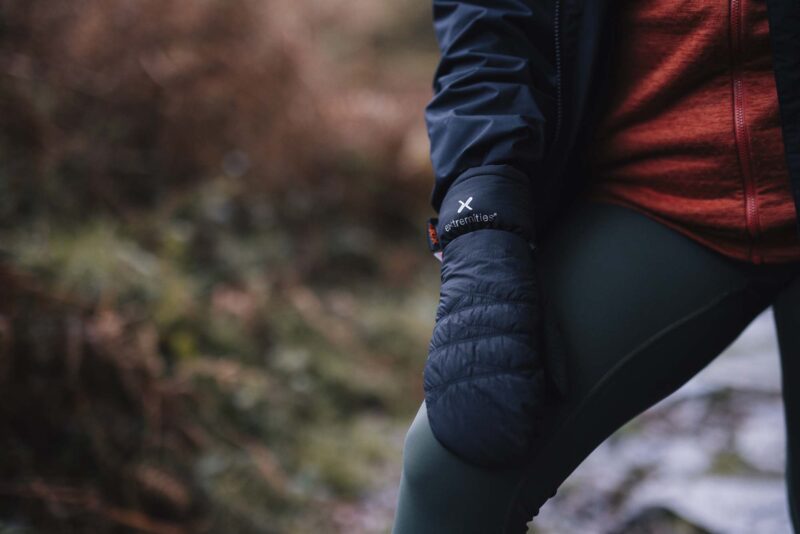 Paradox-Waterproof-Mitts-16-3.jpg Close-up of a person outdoors wearing navy quilted mittens with an "extremities" logo, a red zip-up jacket and dark leggings with one knee bent against a blurred woodland background.