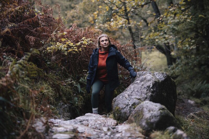 Paradox-Waterproof-Mitts-26-3.jpg Woman in a dark waterproof jacket and orange fleece climbing over mossy rocks in an autumnal woodland, steadying herself with a gloved hand amid ferns and trees.