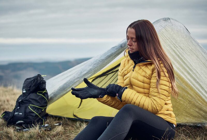 Paradox-gloves-3.jpg A woman in a yellow puffer jacket and black leggings sits by a small yellow-and-silver tent on a grassy hillside, putting on black gloves with a black backpack beside her and cloudy hills in the background.