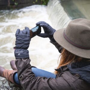 Person wearing a brown felt hat, quilted black gloves and a dark jacket sits on a riverbank edge, holding a smartphone horizontally to photograph a small weir-like waterfall below.