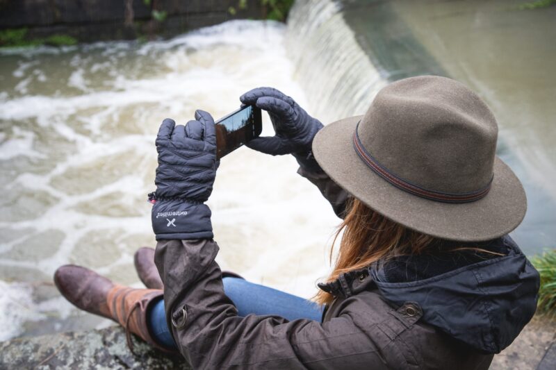 Person wearing a brown felt hat, quilted black gloves and a dark jacket sits on a riverbank edge, holding a smartphone horizontally to photograph a small weir-like waterfall below.