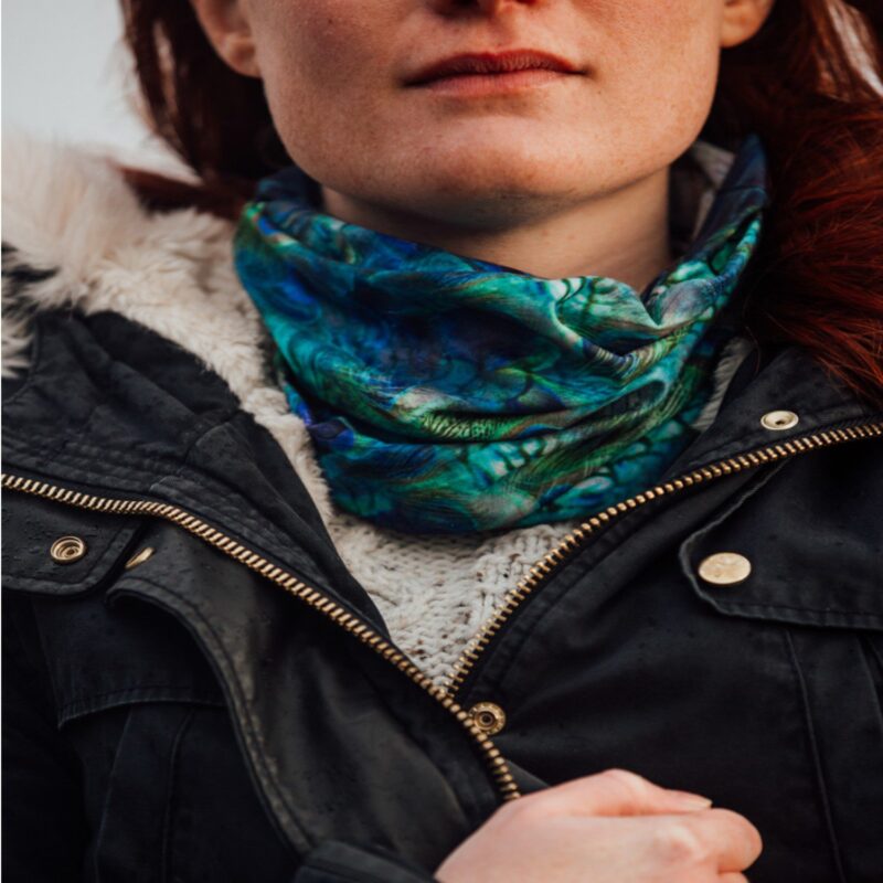 Close-up of a woman's lower face and neck wearing a blue-green patterned neck gaiter over a cream knitted jumper, a dark fur-trimmed jacket with a brass zipper and a hand gripping the jacket.
