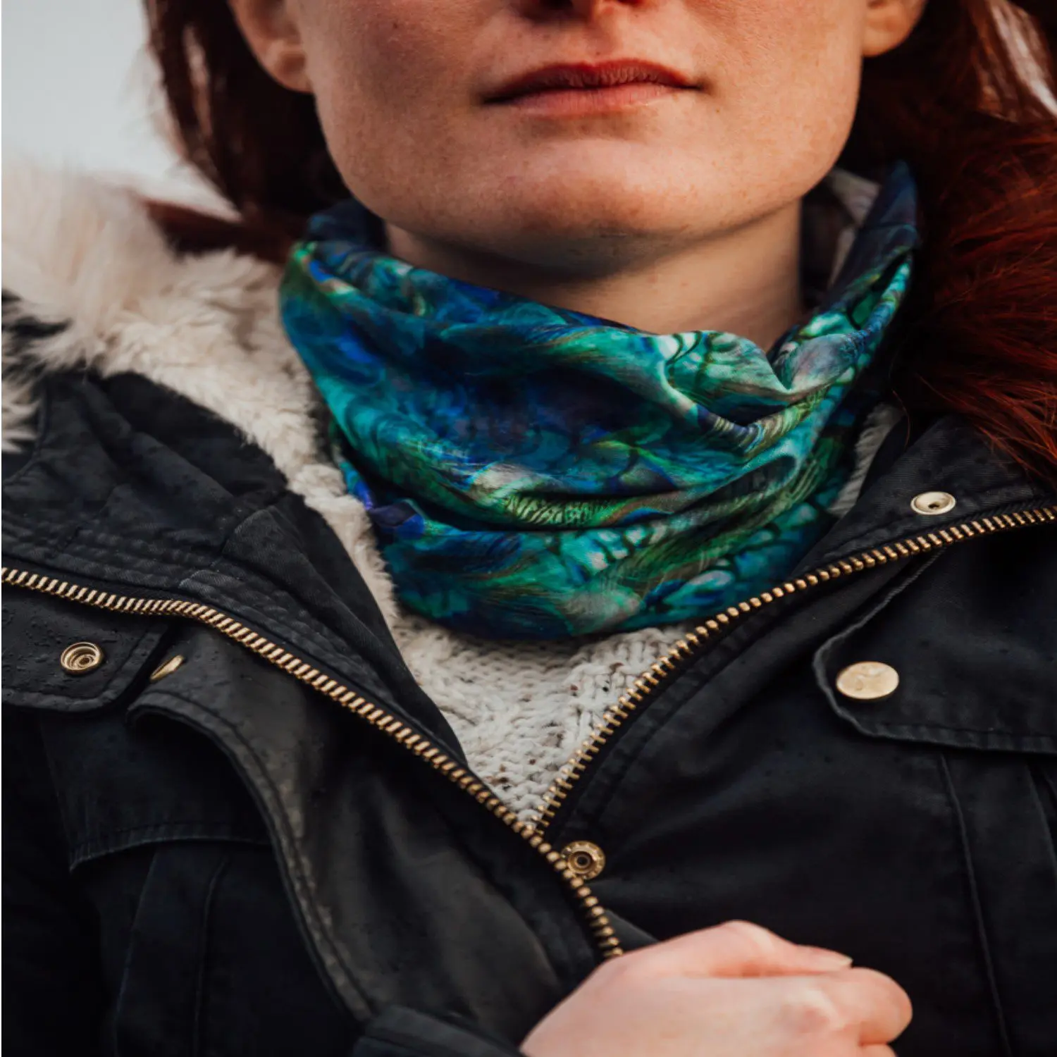 Close-up of a woman's lower face and neck wearing a blue-green patterned neck gaiter over a cream knitted jumper, a dark fur-trimmed jacket with a brass zipper and a hand gripping the jacket.