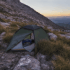 Pioneer-2-in-sunset-e1627480295943-3.png Green two-person tent partially open to reveal sleeping bags, pitched among rocks and tussock grass on a steep mountain slope at sunset with distant layered hills on the horizon.