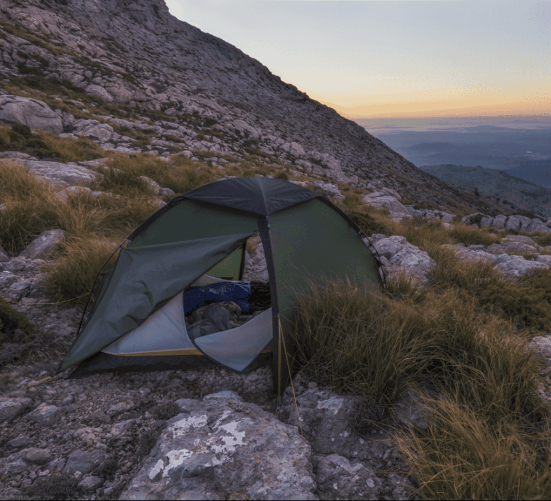 Green two-person tent partially open to reveal sleeping bags, pitched among rocks and tussock grass on a steep mountain slope at sunset with distant layered hills on the horizon.